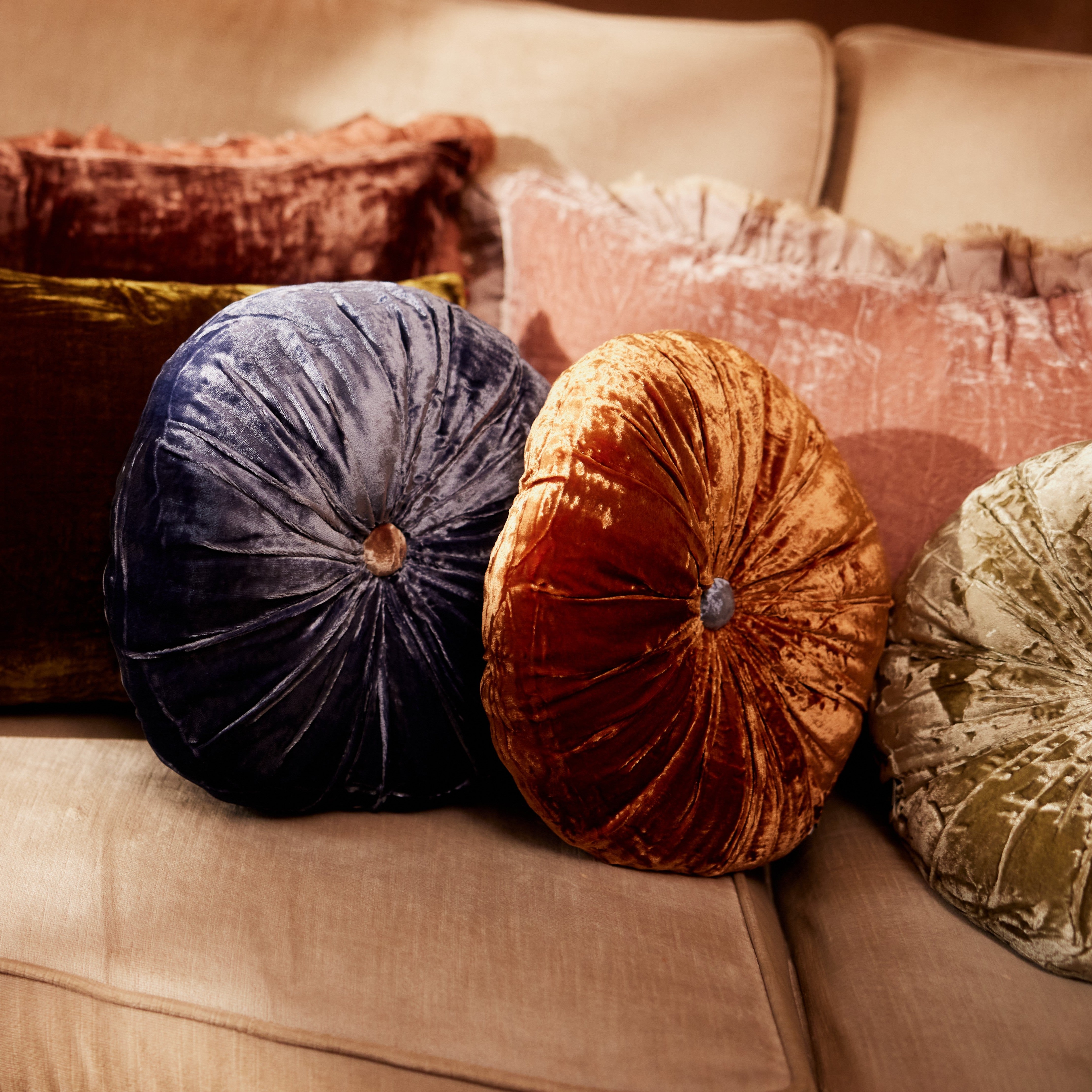 A round silk velvet cushion in delft blue with a contrasting button, displayed with other silk velvet cushions.