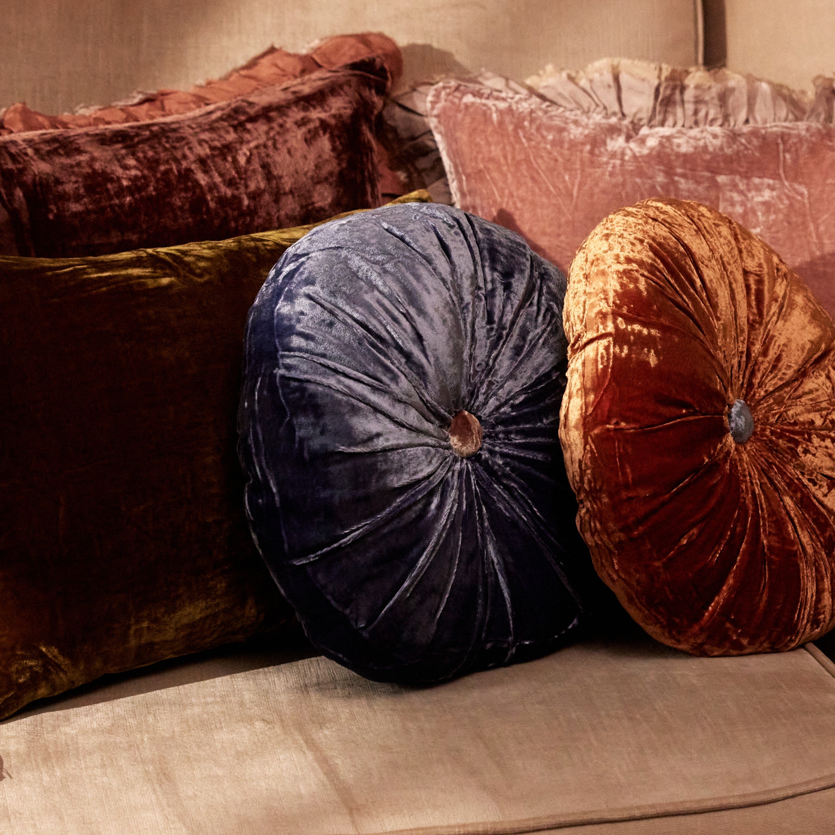 A round silk velvet cushion in delft blue with a contrasting button, displayed with other cushions.