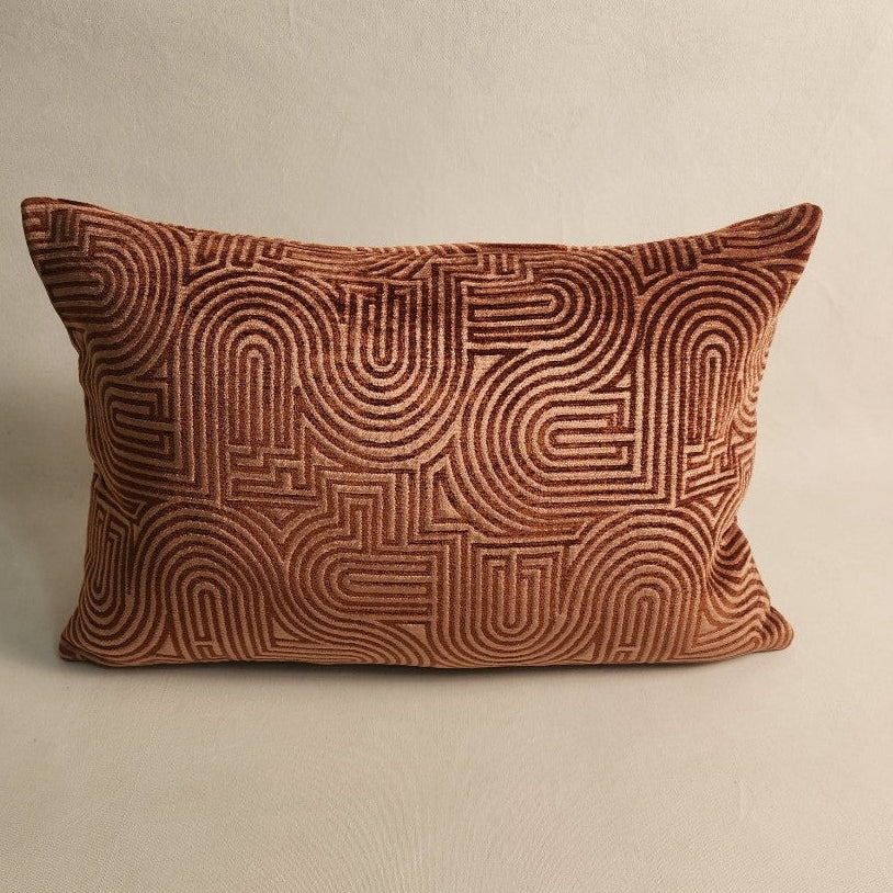 A rectangular throw pillow in rust-brown velvet with an embossed geometric maze-like pattern, set against a neutral beige background.