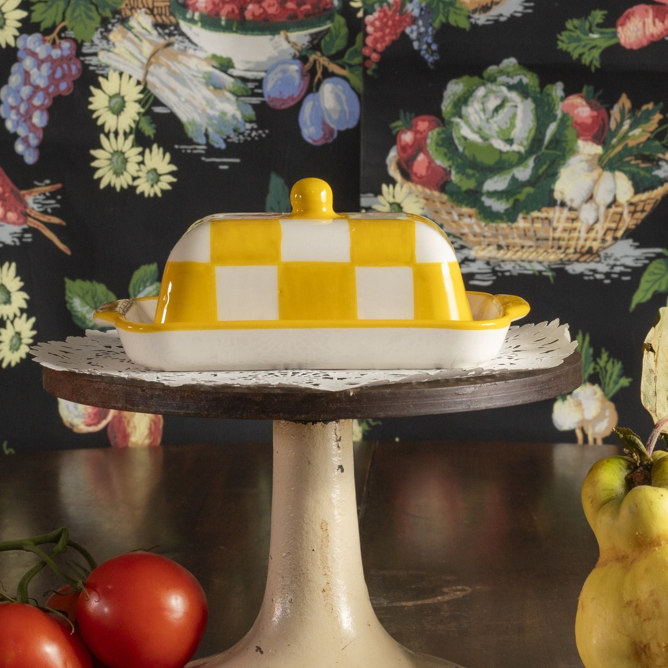Yellow and white checkered butter dish on a wooden stand with a floral and fruit patterned background.