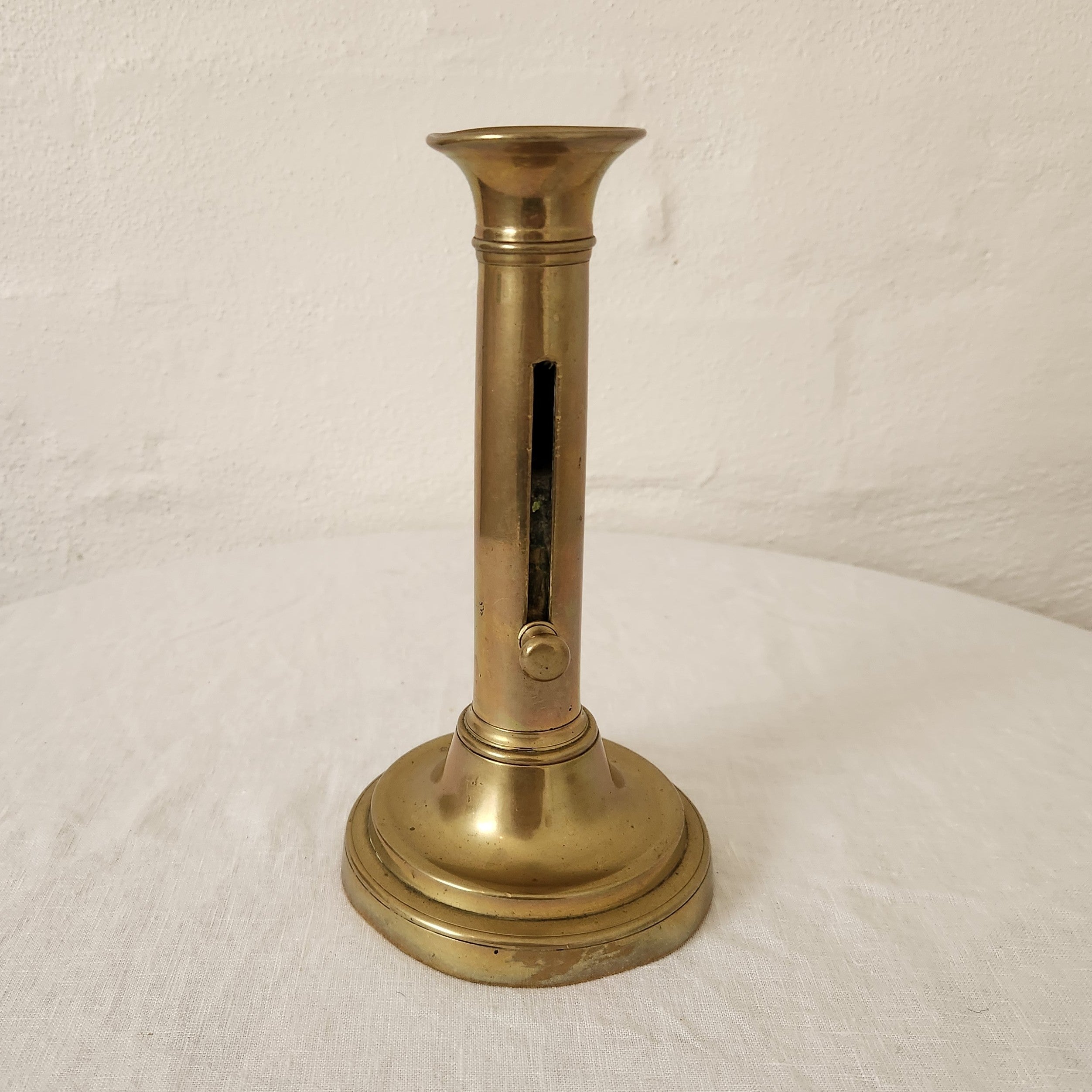 A vintage brass candlestick holder with a circular tiered base and a slender stem featuring a vertical slot and small round detail, set against a white textured wall and white fabric surface.