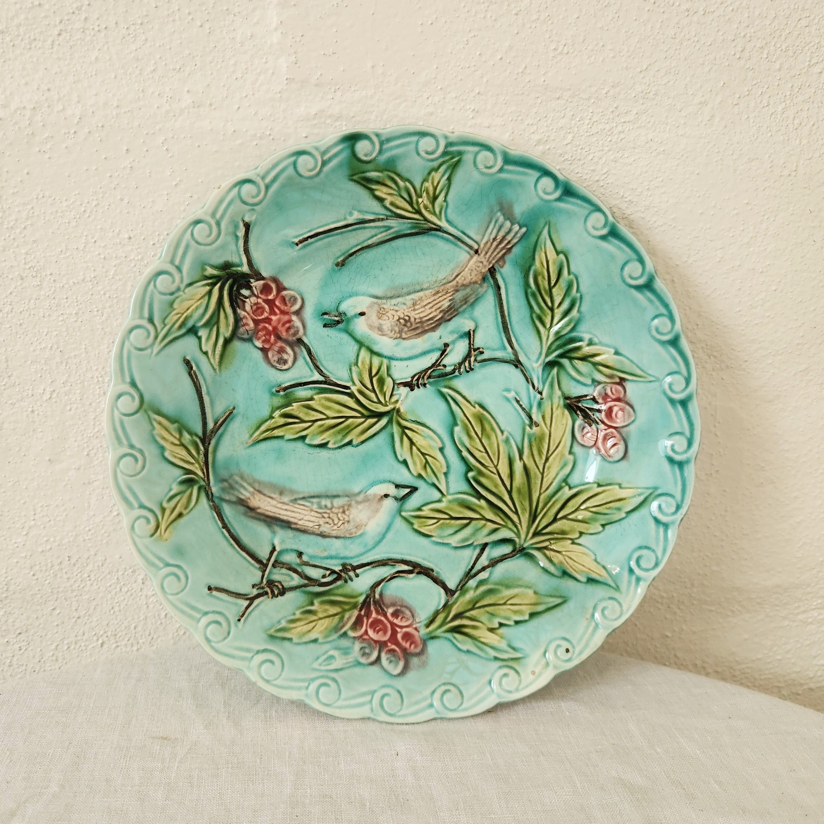 A round turquoise ceramic plate with a scalloped edge, decorated with hand-painted birds, green leaves, and pink berries, set against a textured off-white background.