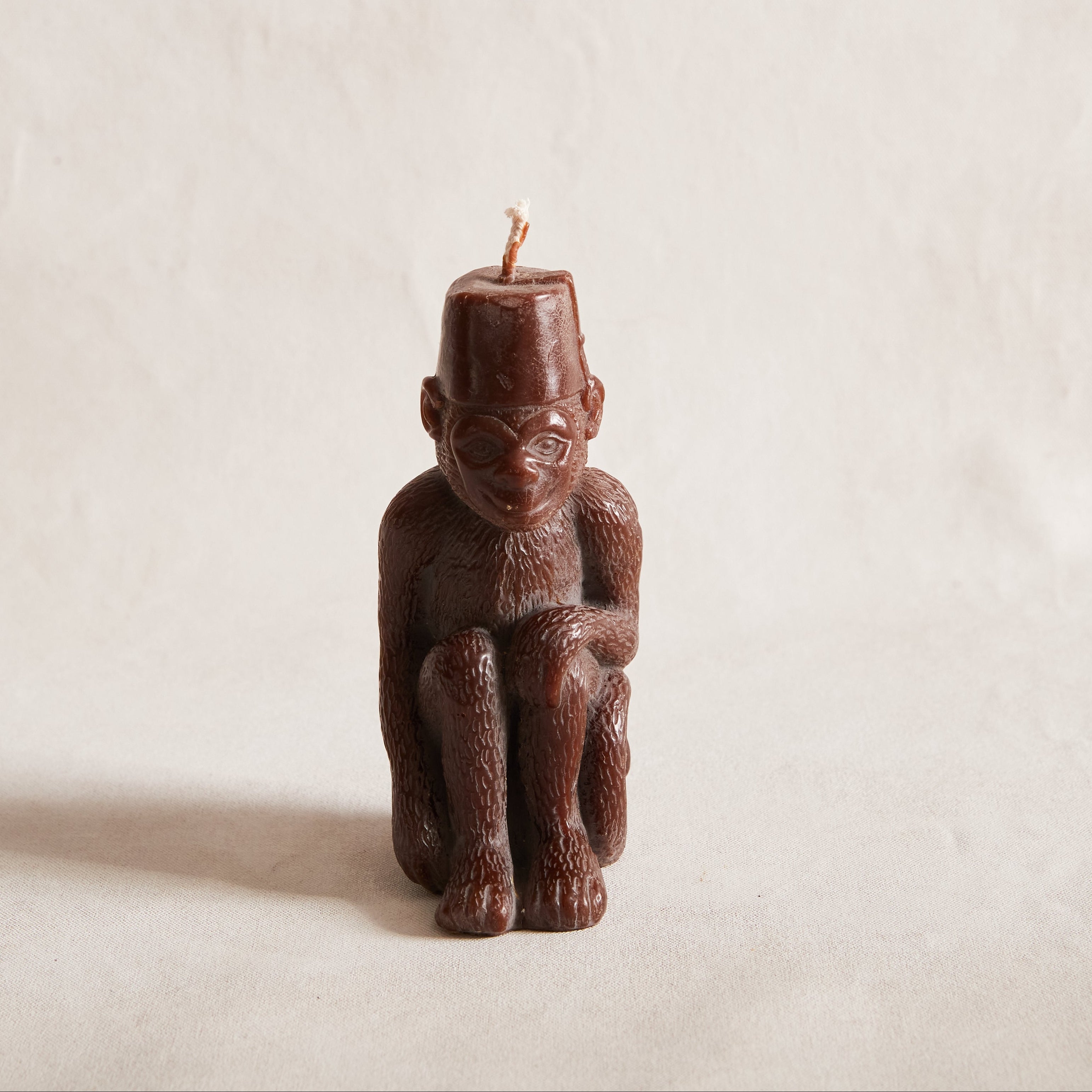 A brown candle with a monkey figurine design, sitting with a fez on its head, set against a neutral background.