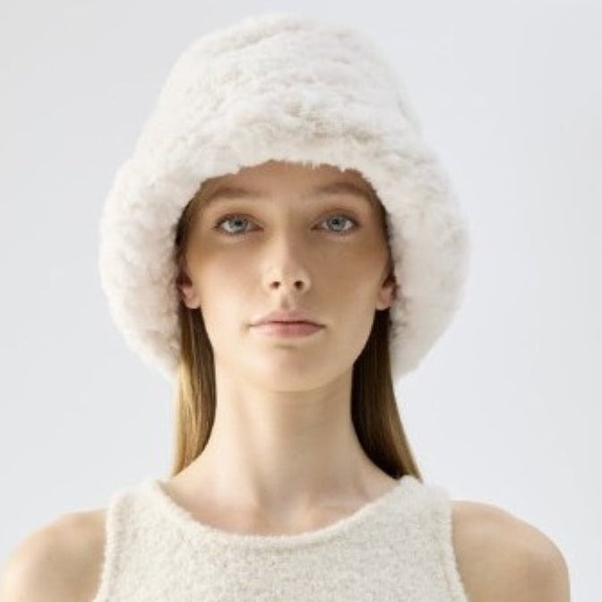 A woman wearing a fluffy cream-colored bucket hat and a matching textured sleeveless top, photographed against a clean light gray studio background.