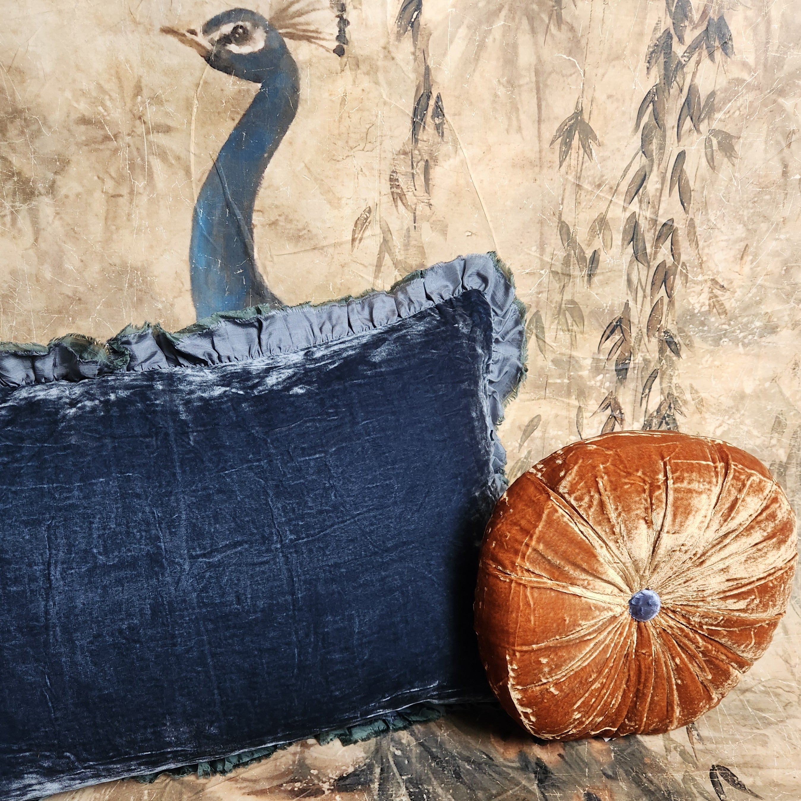 A round silk velvet cushion in Bombay orange with a contrasting button, displayed with another silk velvet cushion on a peacock background