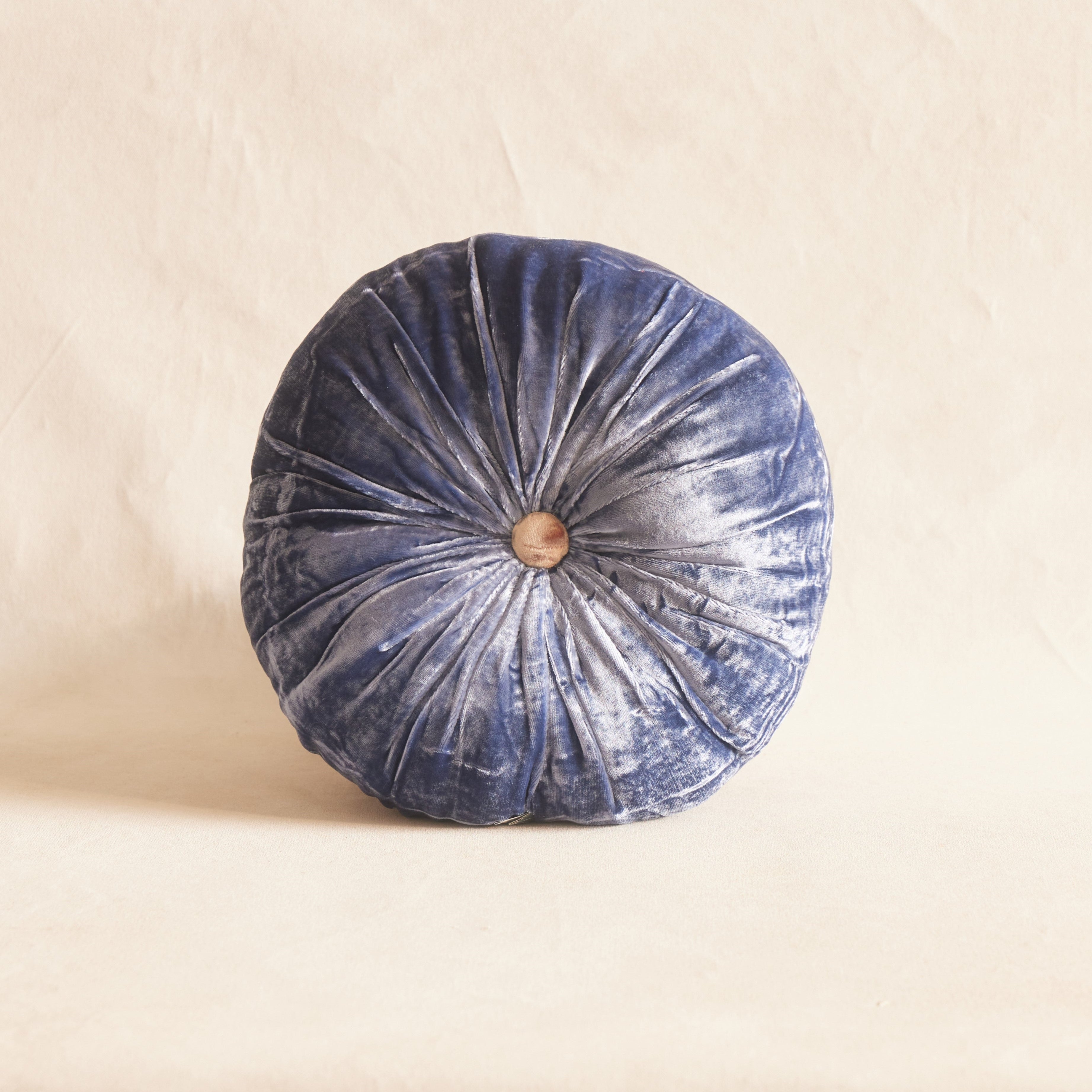 A round silk velvet cushion in delft blue with a contrasting button, placed on a neutral background.