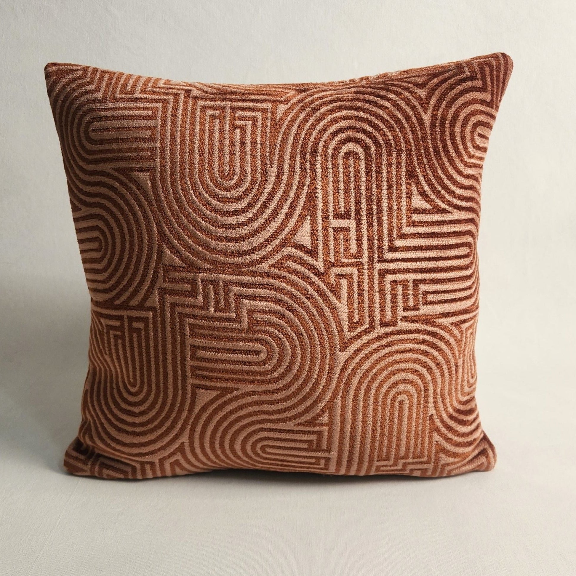 A square throw pillow in textured rust-brown velvet with a raised geometric maze-like pattern, set against a neutral off-white background.