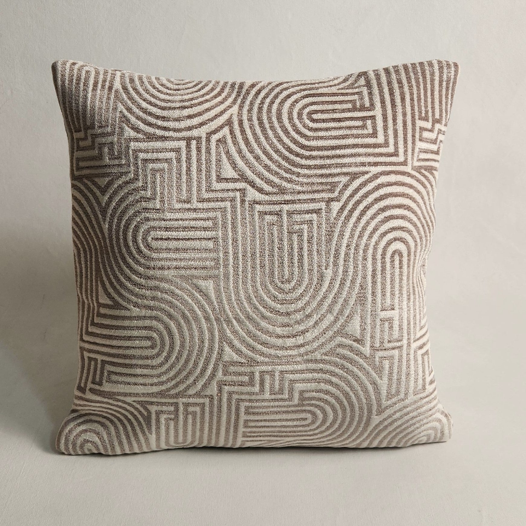 A square throw pillow featuring a textured geometric pattern of concentric arches and maze-like lines in taupe and light beige, set against a neutral light gray background.