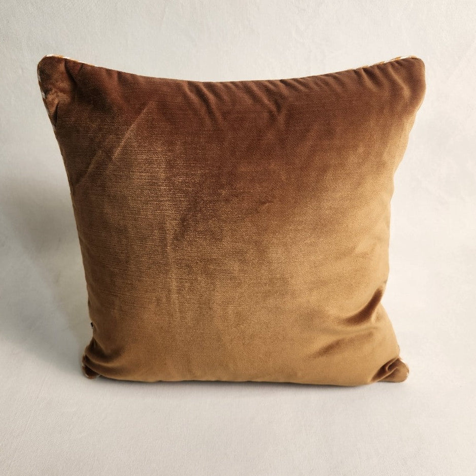 A square velvet throw pillow in a warm camel brown color, photographed against a neutral, textured off-white background.