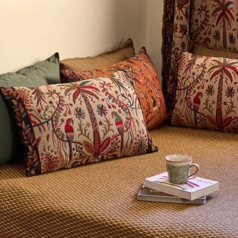 A styled indoor seating area with woven jute cushions and matching tropical bird-and-floral prints. The scene includes multiple decorative pillows and a curtain, with a small stack of books and a patterned mug on the seat.