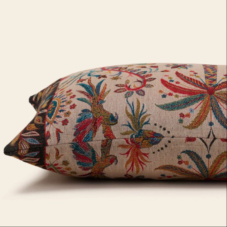 A close-up of a decorative throw pillow featuring a vibrant, multicolored embroidered pattern of birds and floral motifs on a beige linen-like fabric, set against a solid cream background.